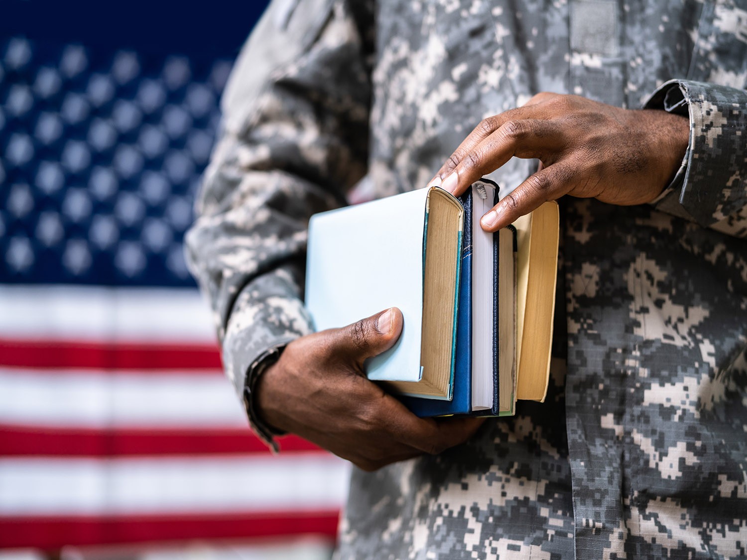 Serviceman holding textbooks standing in front of American flag.