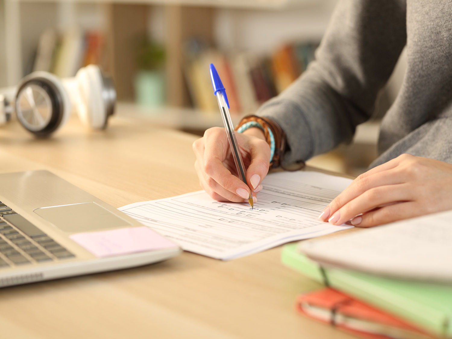 Hands filling out paperwork surrounded by school books, laptop and headphones